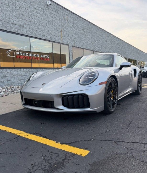 Silver sports car parked outside an auto shop with a gray building and yellow parking line.