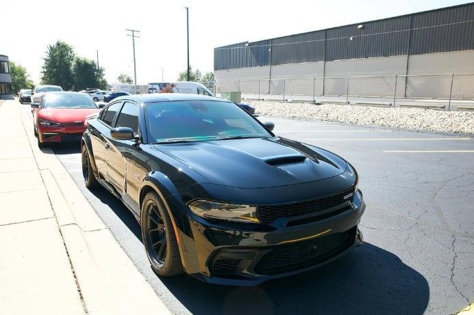 Black sports car parked on a sunny street with a red car behind it and a warehouse wall nearby