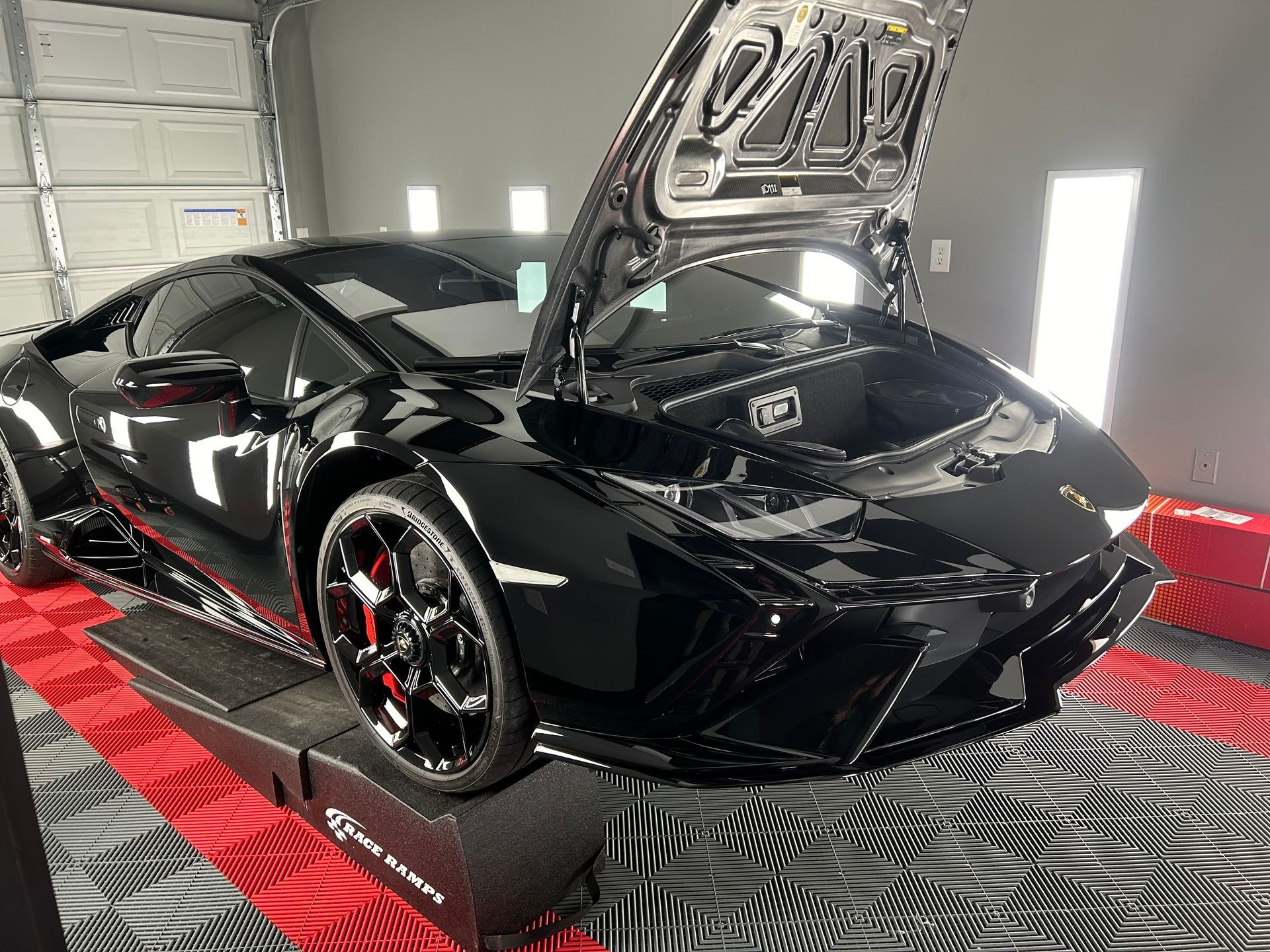Black Lamborghini Huracan with hood open in a well-lit garage, parked on a red and black tiled surface.