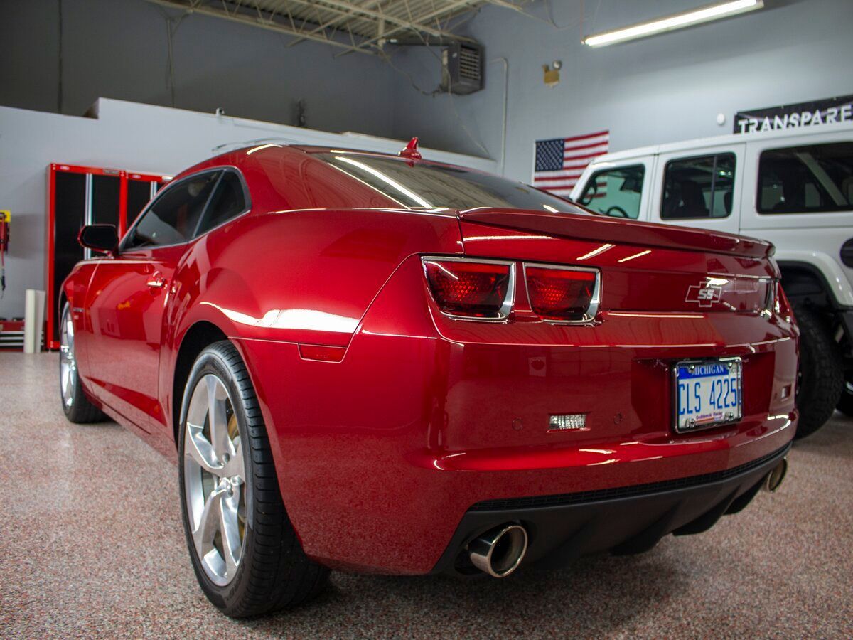 Red Chevrolet Camaro parked inside a garage, next to a white Jeep, with a US flag.