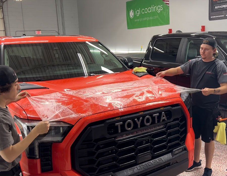 Two people applying protective film to a red Toyota truck hood in a garage.