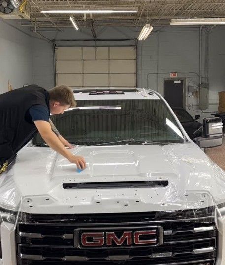 A technician in a garage applies a clear protective film to the white hood of a GMC truck.