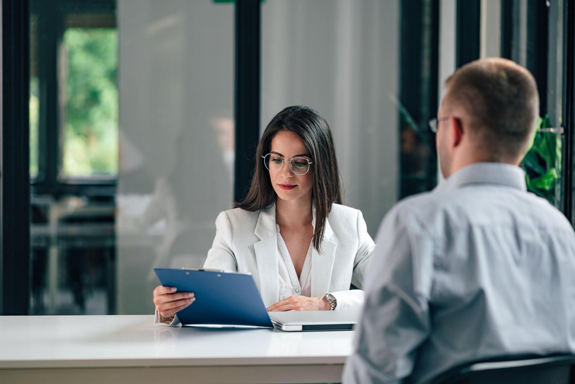 A professional woman in a white blazer reviews a blue folder while seated at a table across from a colleague.