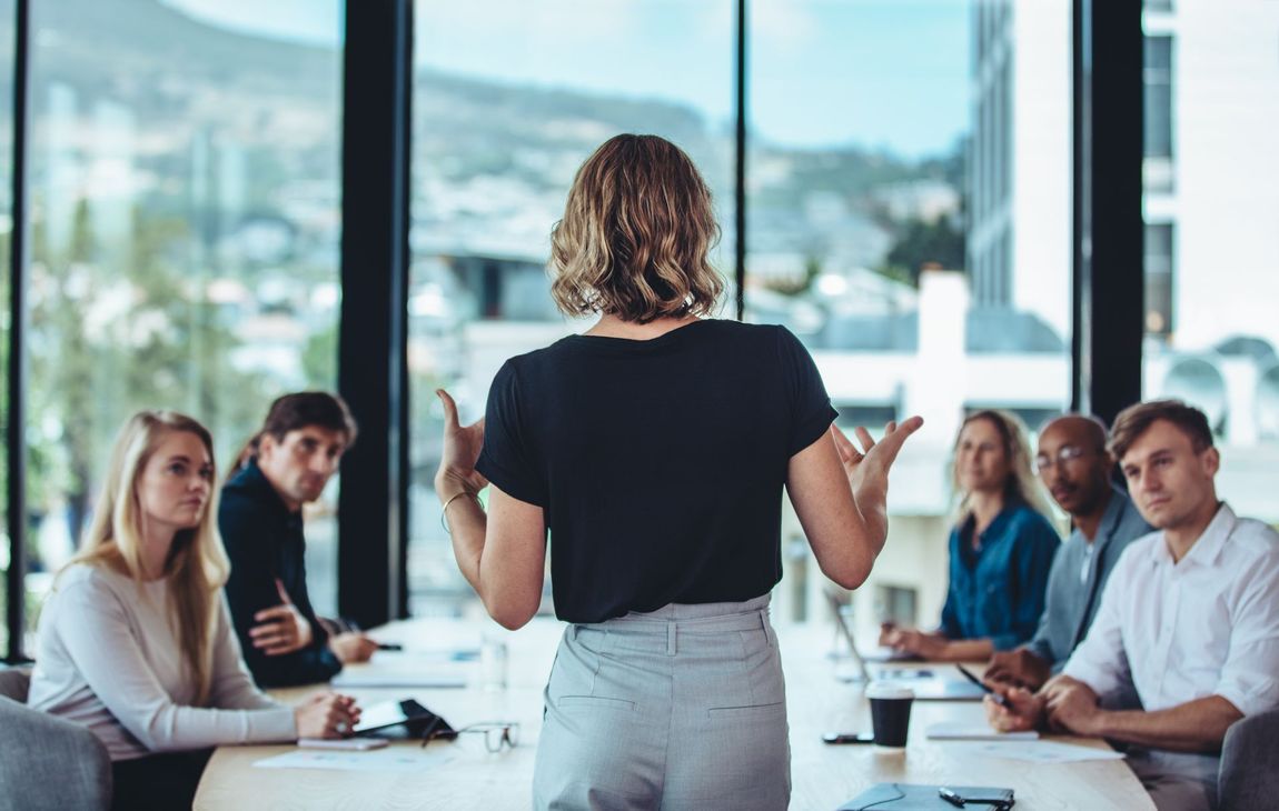 A person stands at the head of a table giving a presentation to a group of colleagues.