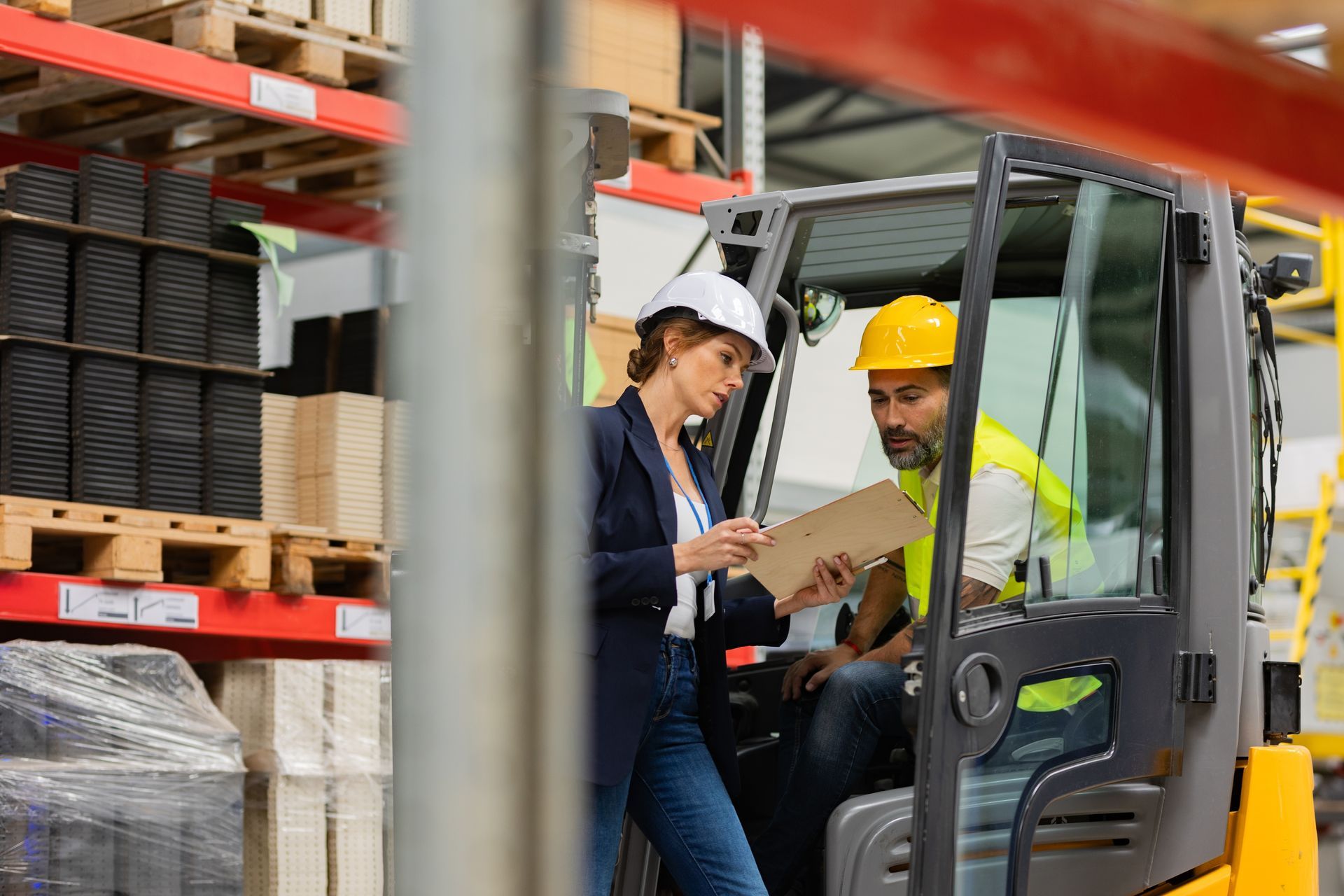 A person in a warehouse setting reviews a document with a forklift operator sitting in their vehicle.