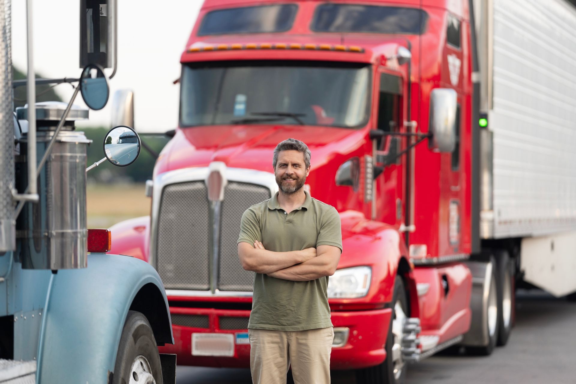 A person with crossed arms stands smiling in front of a red semi-truck.