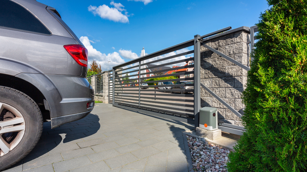 Gray SUV beside a modern metal driveway gate and stone wall under a blue sky
