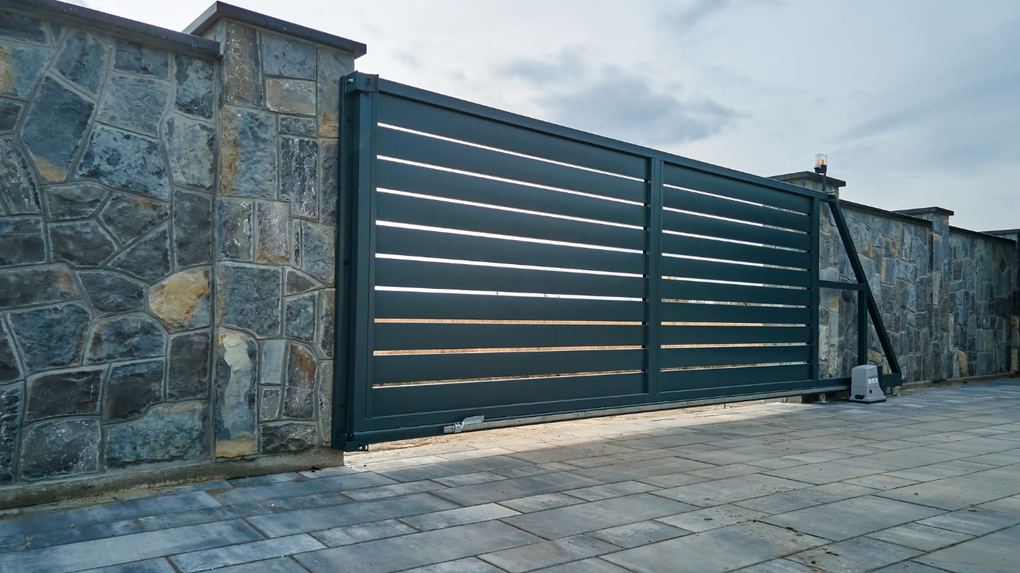 Gray stone wall with a dark green sliding gate on a paved driveway under an overcast sky