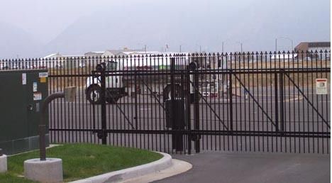 Black metal security gate in front of a fenced industrial lot.