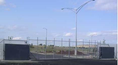 Empty fenced lot with chain-link gate, tall streetlights, and a blue sky overhead