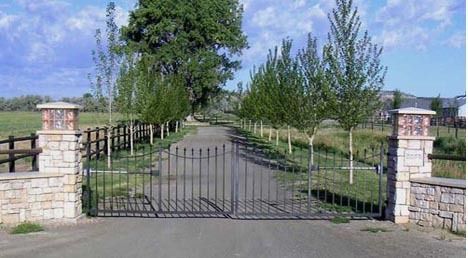 Closed black metal gate at the entrance to a tree-lined driveway, flanked by stone pillars under a blue sky