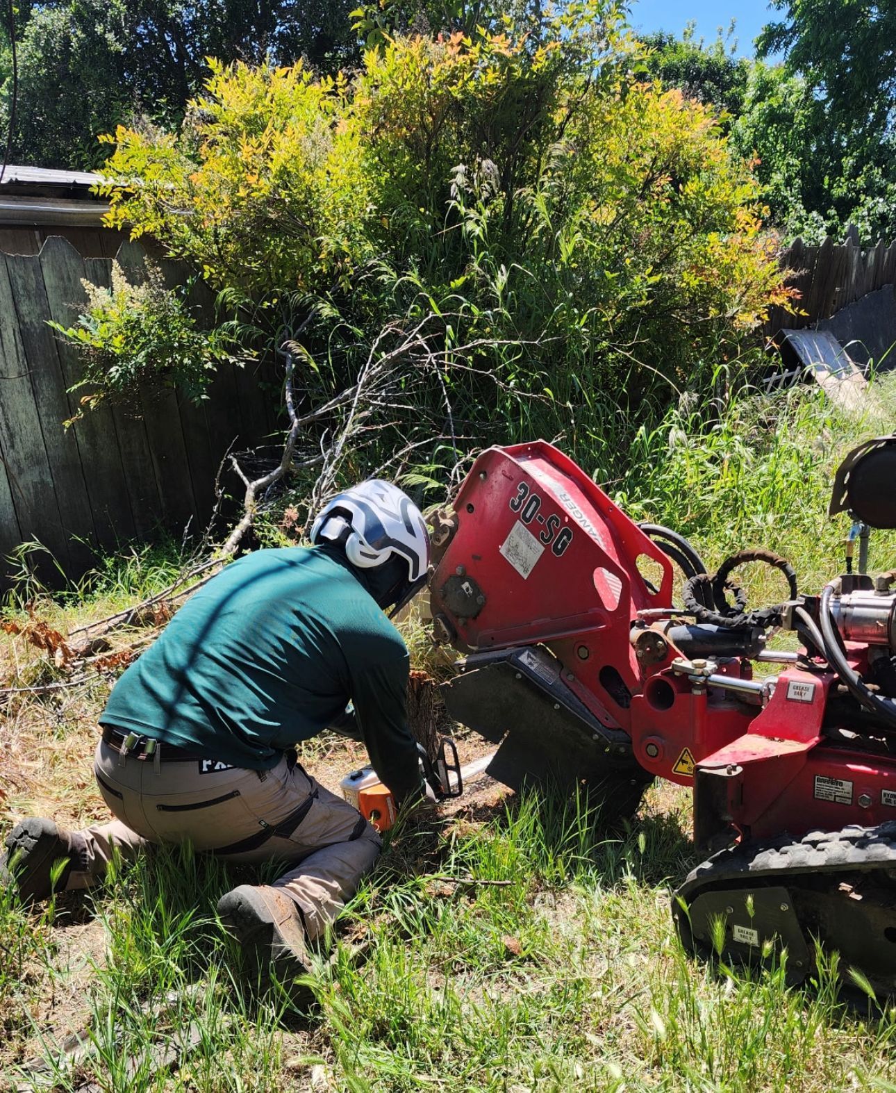 Person kneeling beside a red garden machine in a grassy backyard, wearing a helmet and green shirt