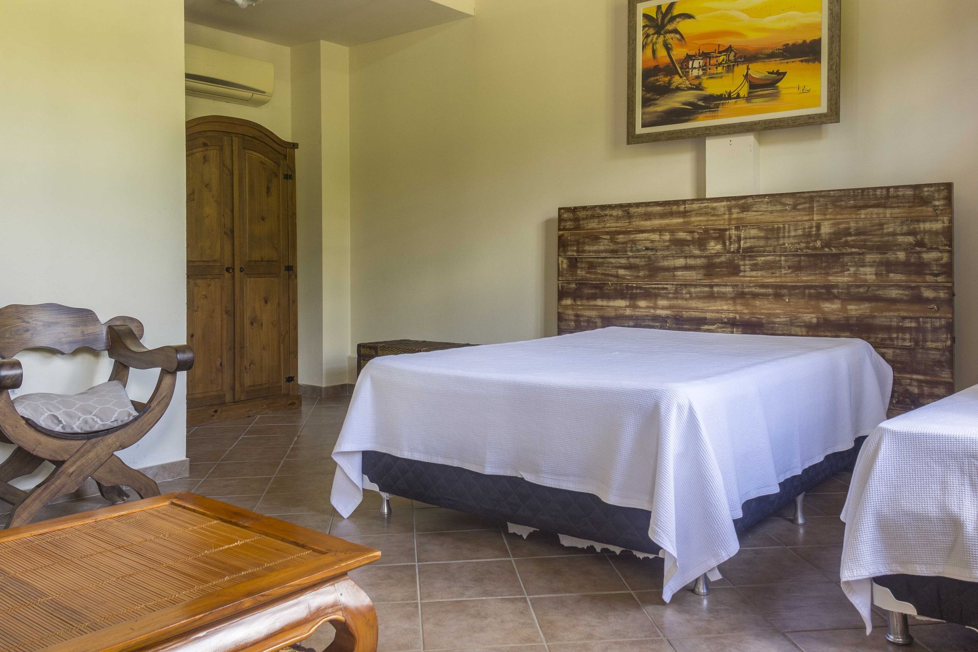 Quarto com duas camas, cabeceira de madeira, cadeira, guarda-roupa e obras de arte emolduradas na parede.