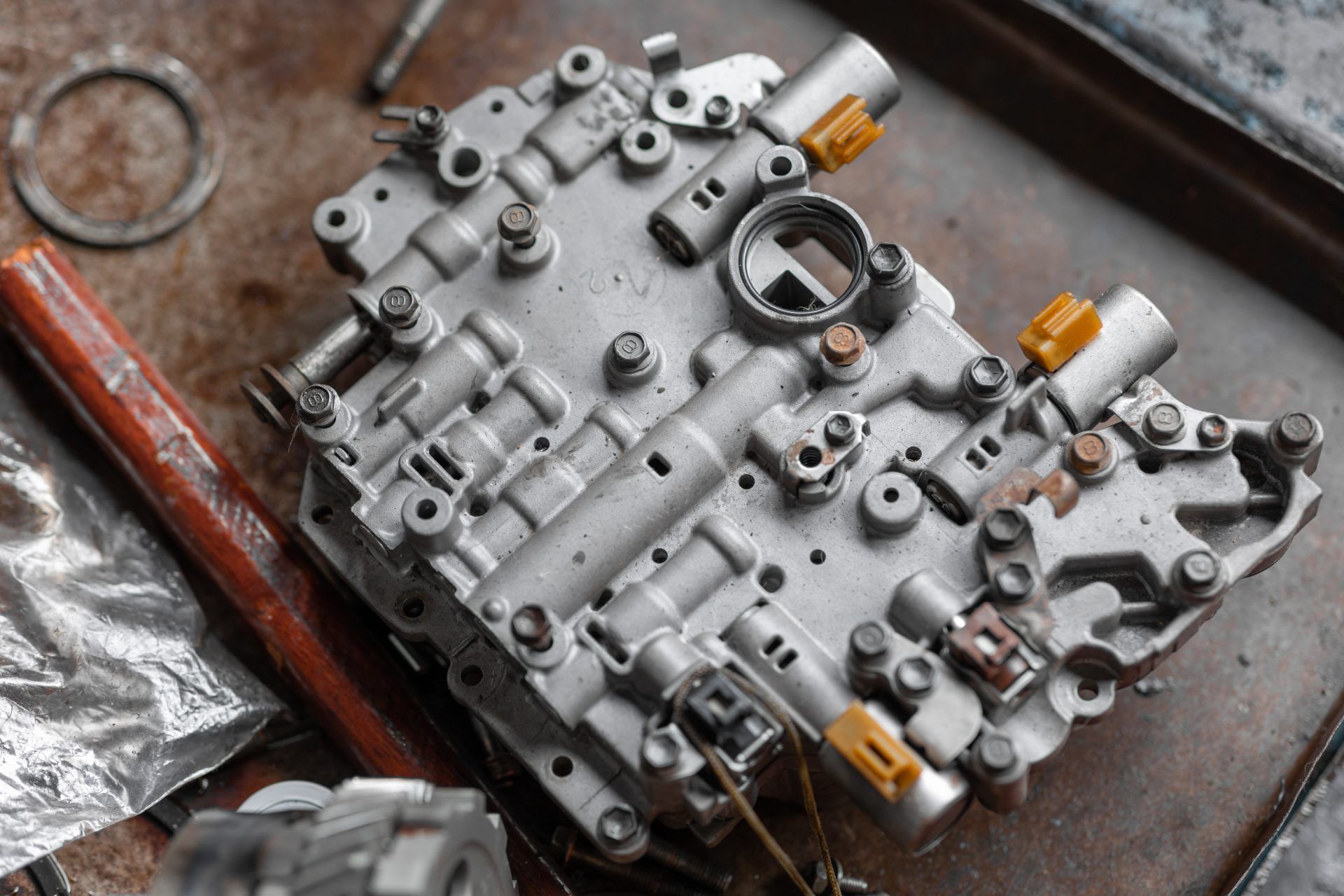 A silver metal automatic transmission valve body with yellow electrical connectors on a dark, industrial surface.
