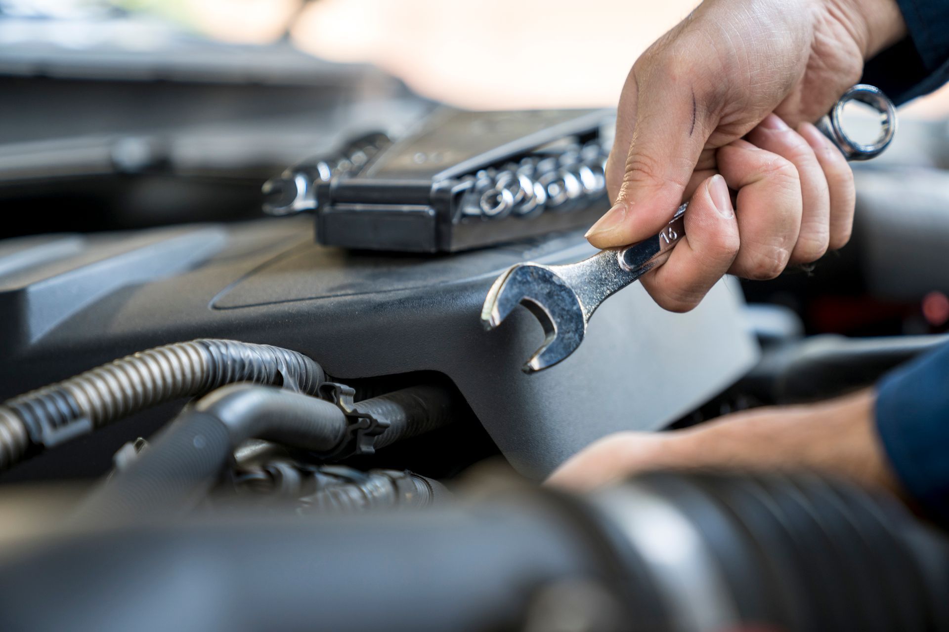 Hands work on a car engine under the hood, holding a metal wrench near a black plastic engine cover.