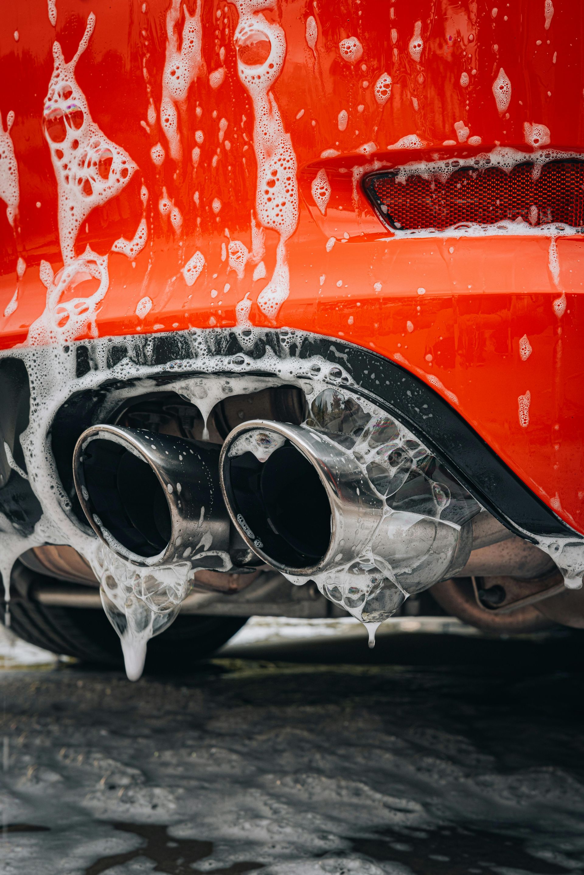 A close-up of a vibrant orange car bumper with dual chrome exhaust pipes covered in white soap suds during a car wash.