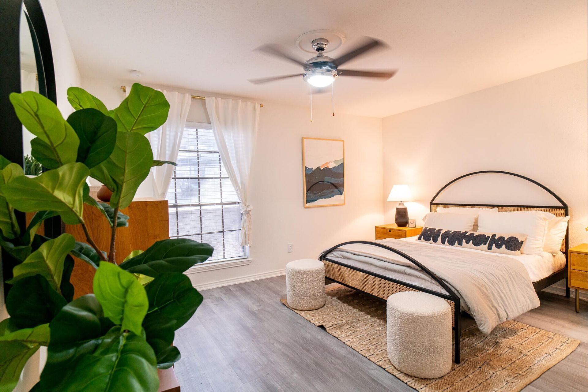 A bright, modern bedroom with light gray floors, a bed with a metal frame, two textured ottomans, and a large plant.