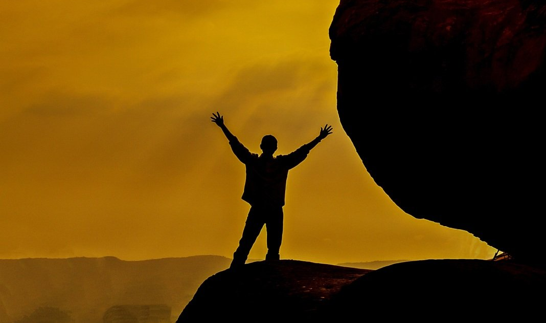 Silhouette einer Person mit erhobenen Armen auf einem Felsvorsprung vor einem goldenen Sonnenuntergang, neben einem großen Felsbrocken
