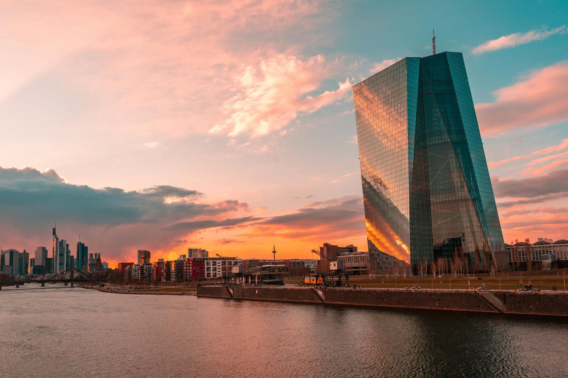 Sonnenuntergang über einem Fluss mit einem modernen Glasturm und der Skyline der Stadt, in der sich warme rosa-orange Wolken spiegeln.