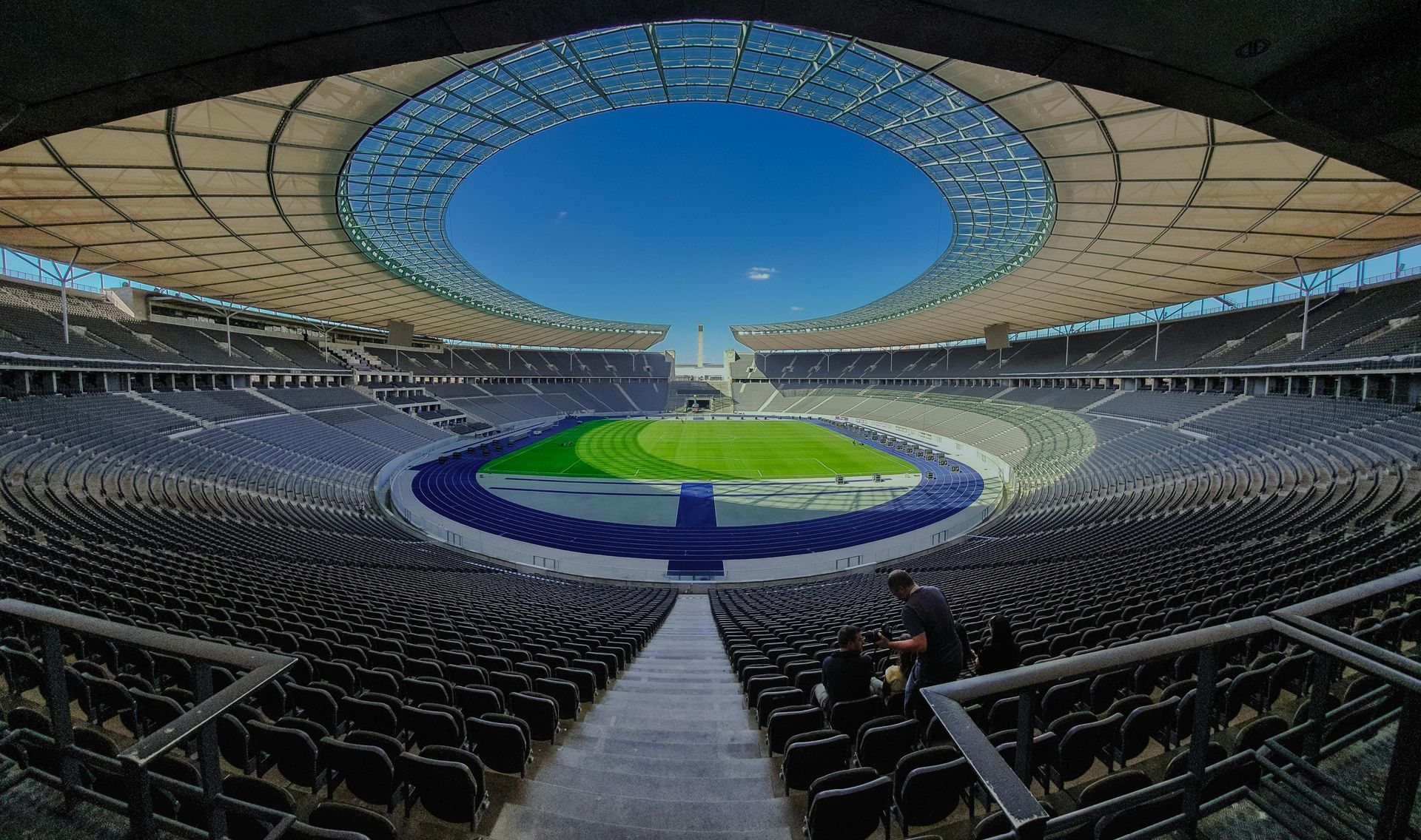 Weitwinkelaufnahme eines modernen Stadions mit rundem Dach, grünem Rasen und blauem Himmel.