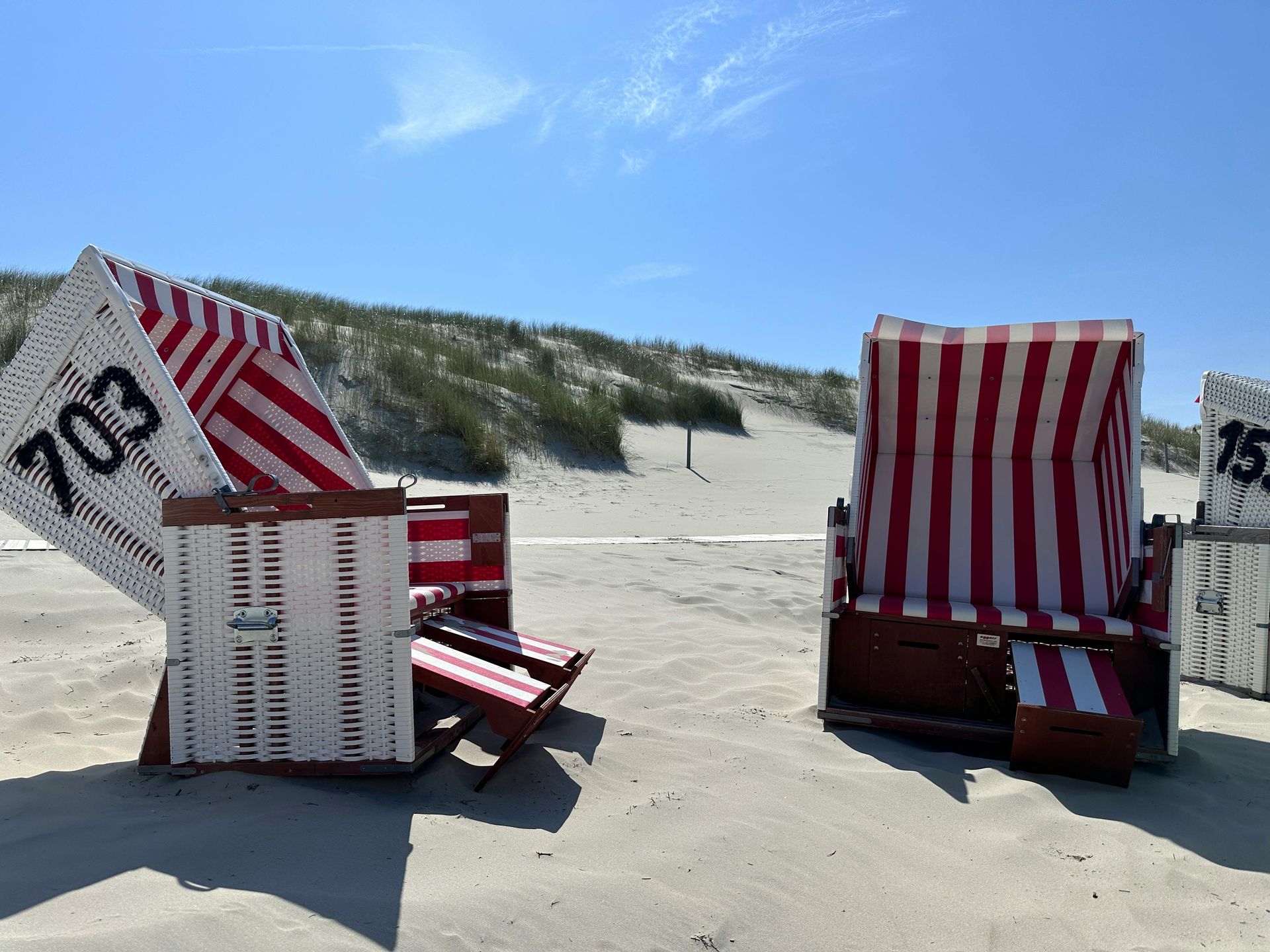 Zwei gestreifte Strandstühle im Sand, im Hintergrund Dünen und blauer Himmel.