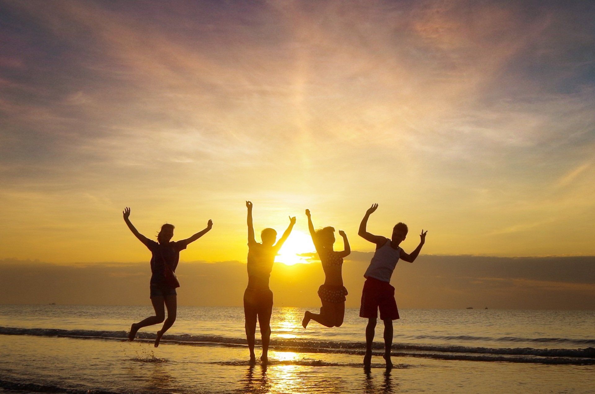 Als Silhouetten zeichnen sich Freunde ab, die bei Sonnenuntergang am Strand springen, während sich die Sonne im Wasser spiegelt.