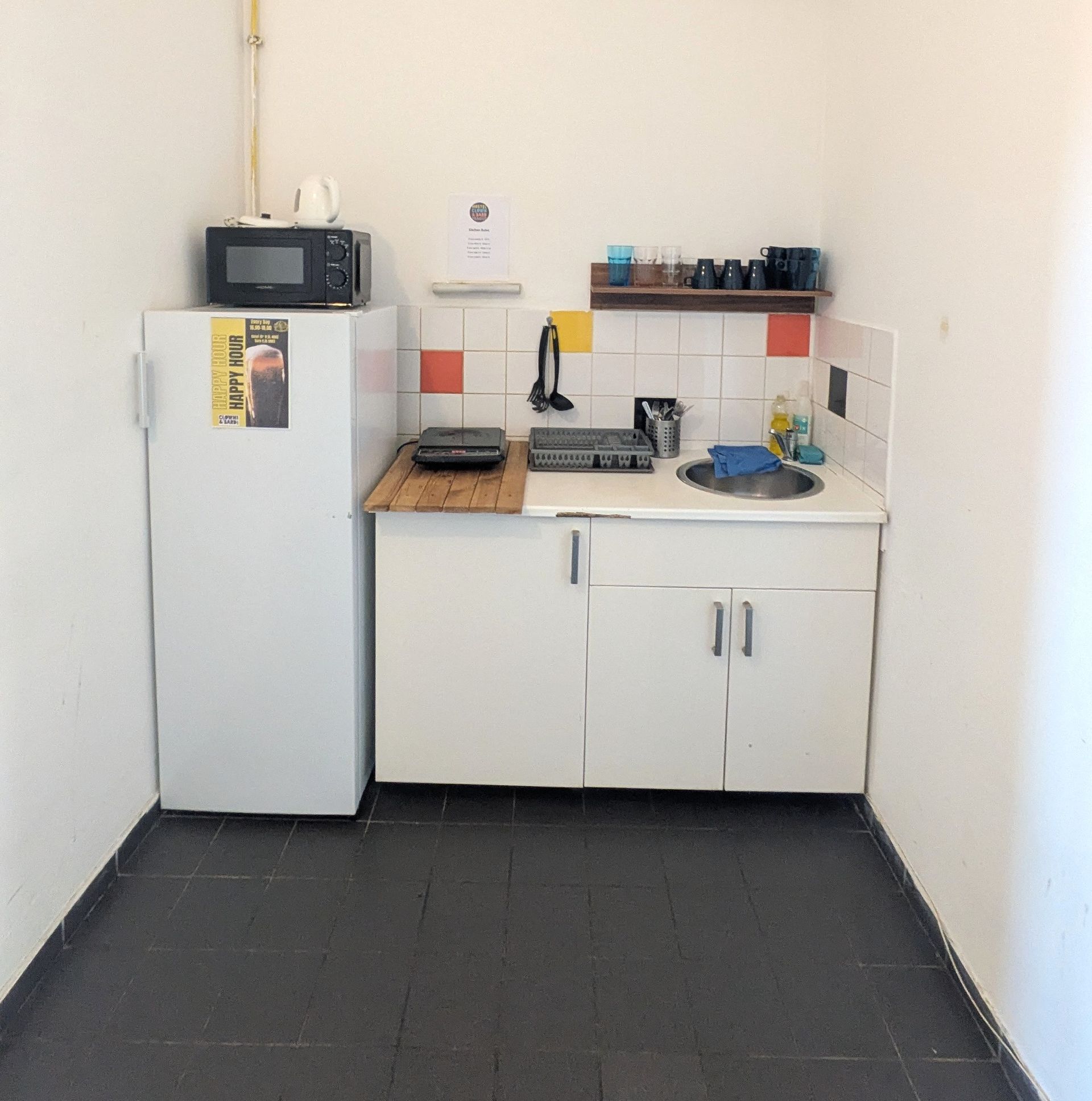 simple kitchen: refrigerator, microwave, sink, cabinets, dishes on shelf, and dark tile floor.
