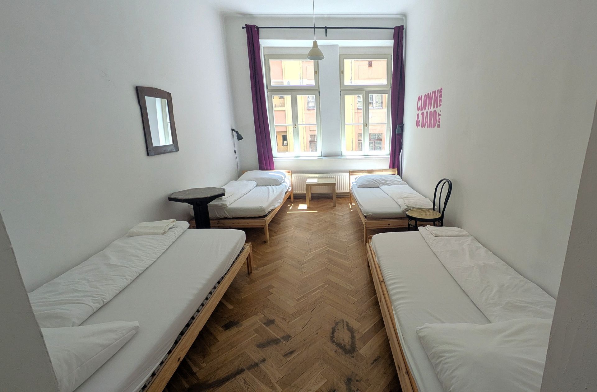 A hostel room with four beds, a small table, a chair, and a window.
