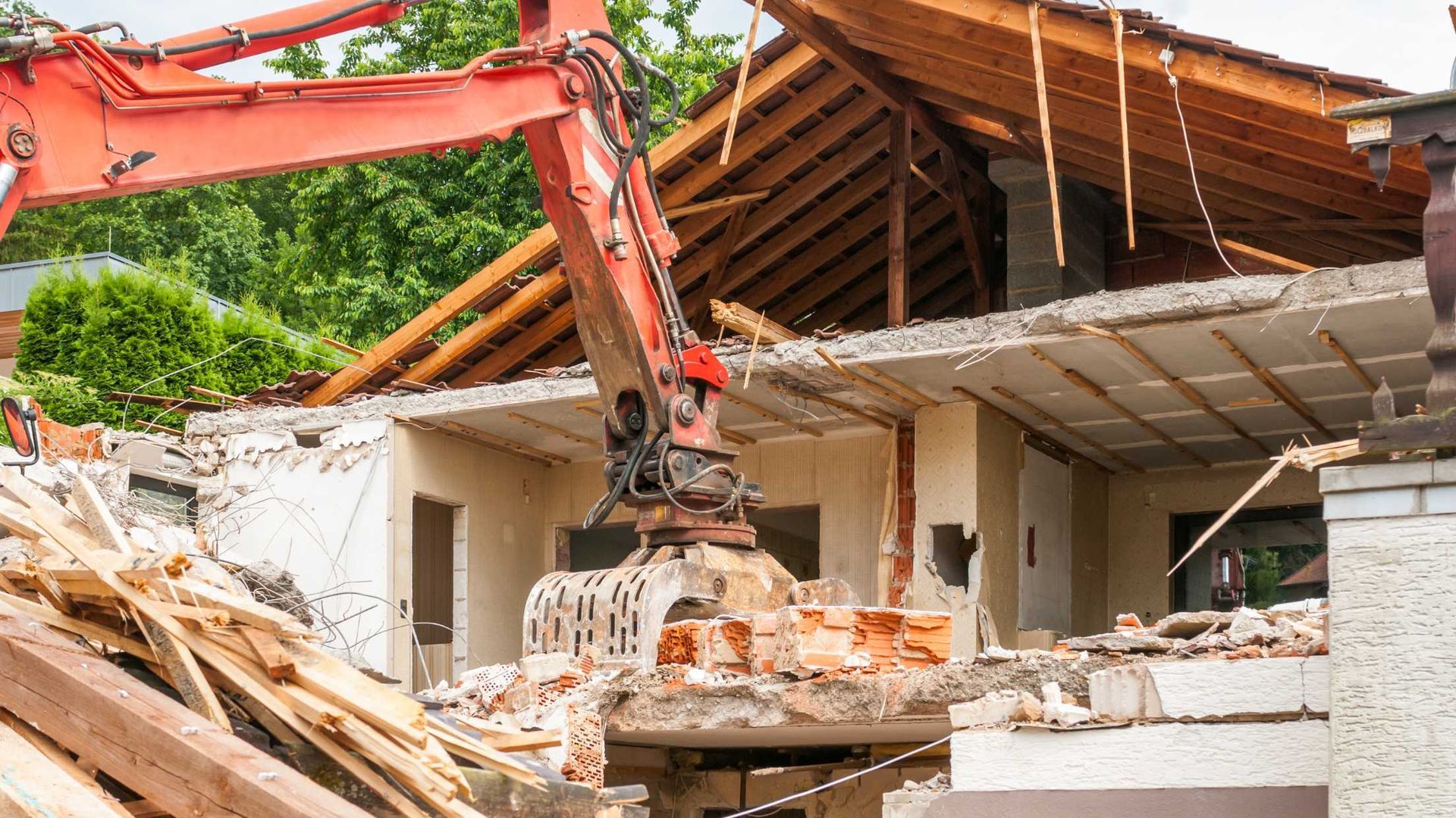 Excavator demolishing a damaged house with broken roof and rubble around it