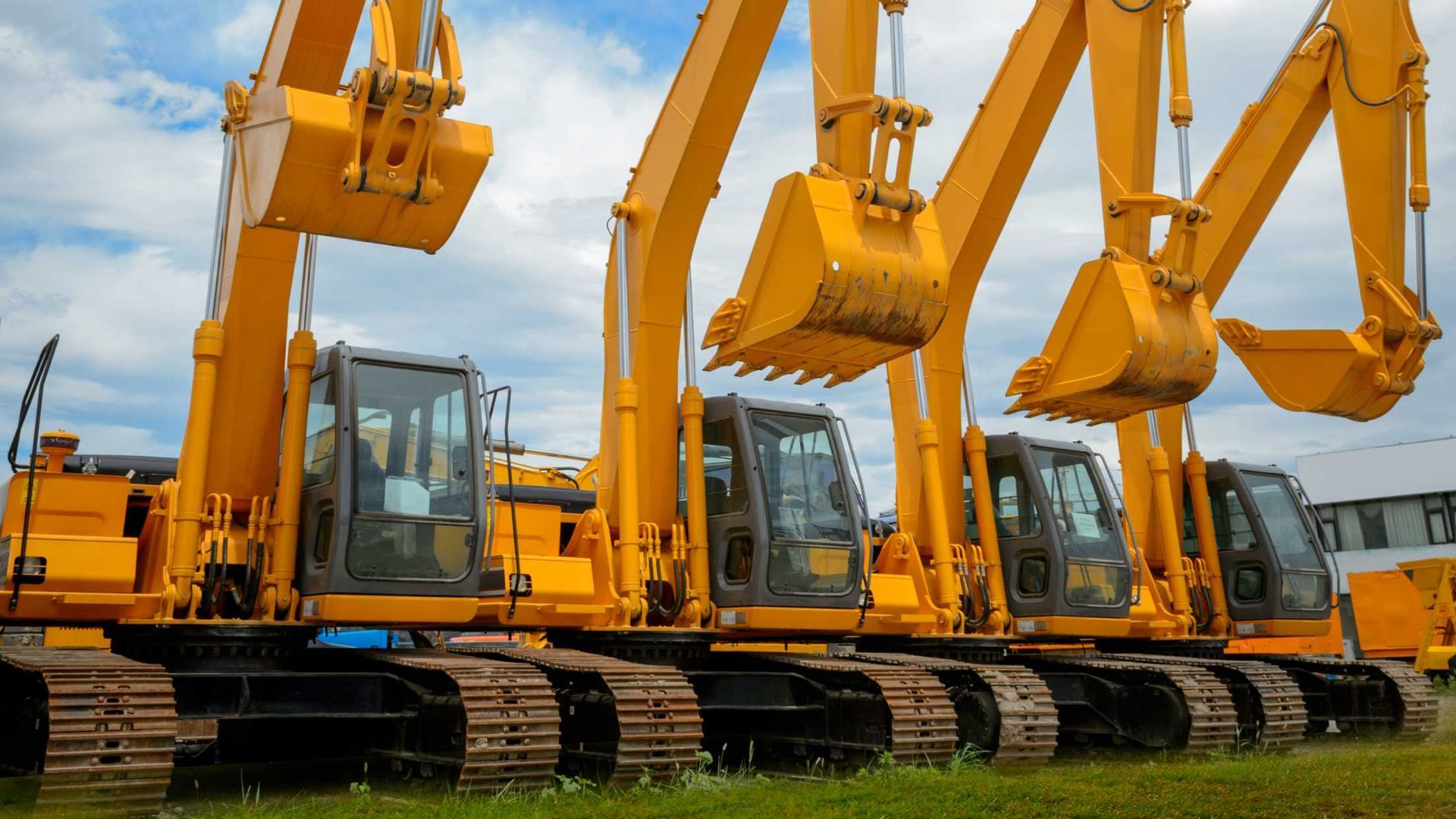 Row of yellow excavators lined up on a construction site with muddy tracks and grass foreground