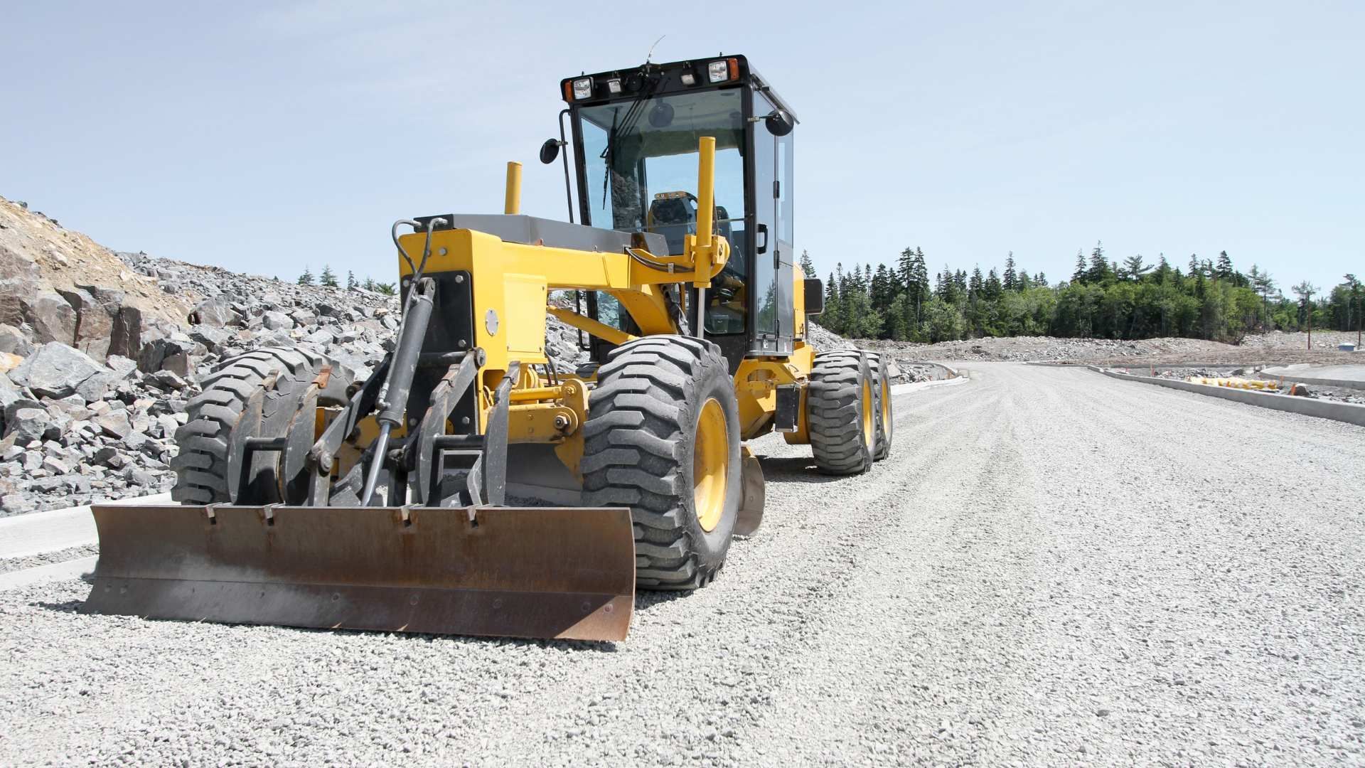 Yellow road grader leveling gravel on a construction site beside a rock pile under a clear sky