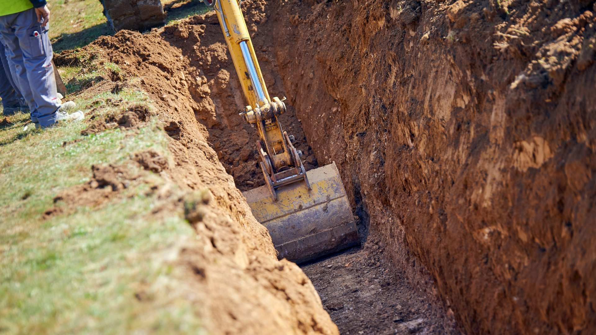 Excavator bucket digging a long trench in a dirt field beside a grassy edge
