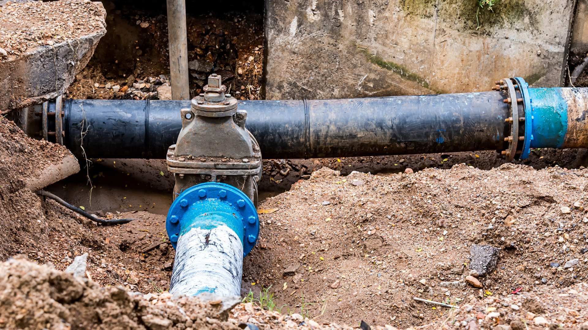 Exposed water main pipes and valve joint in a dirt trench, with blue couplings and black pipe.