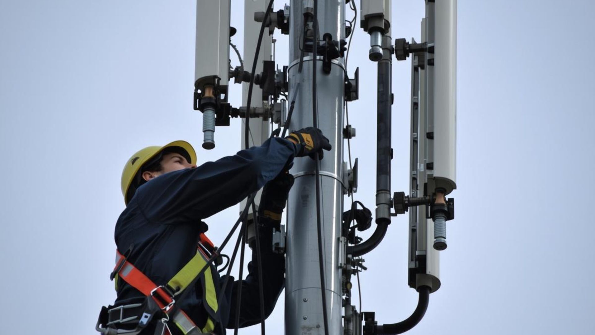 Worker in safety gear climbing a telecom tower, adjusting equipment against a gray sky