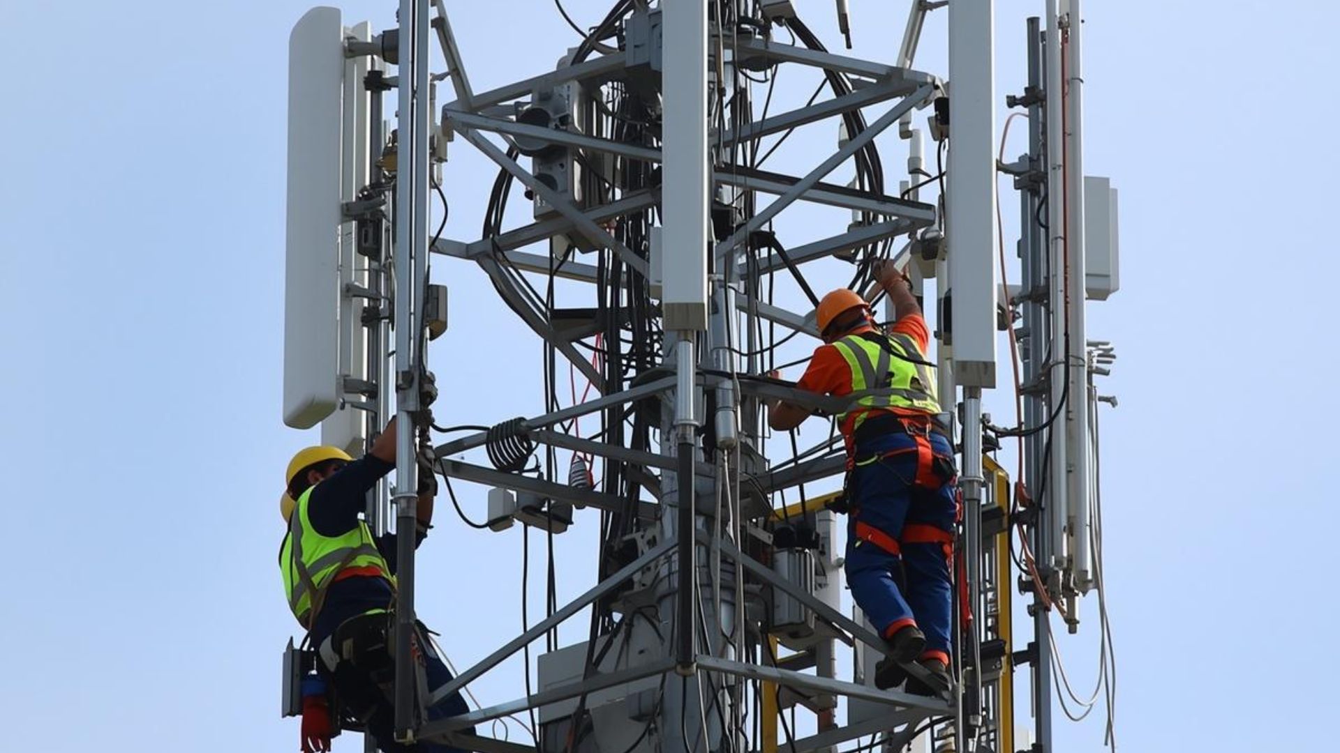 Workers in safety gear climbing a cell tower, adjusting equipment high above ground.