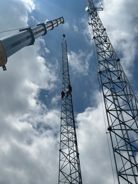 Cell tower with antennas against a bright blue sky, beside a small utility building on a rural plain