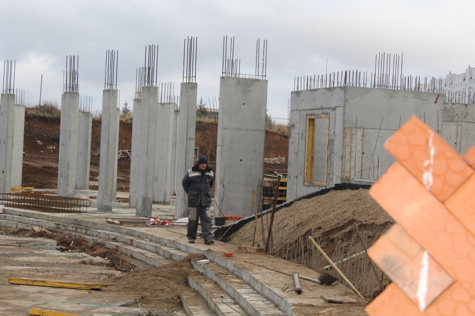 Construction site with concrete columns, exposed rebar, workers, and dirt piles under an overcast sky