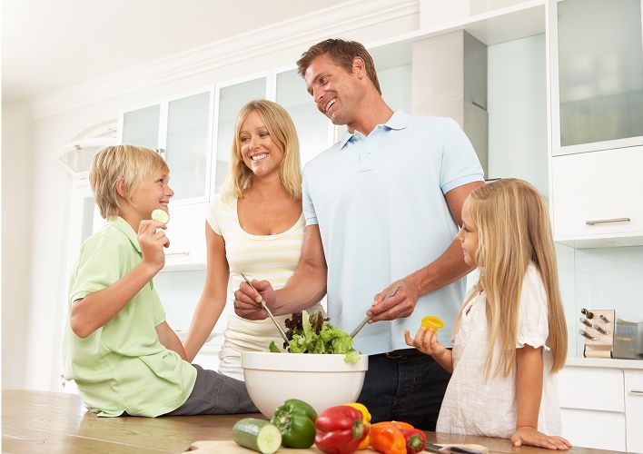 Family preparing food together in a modern kitchen. Mom, dad, son, and daughter smile.