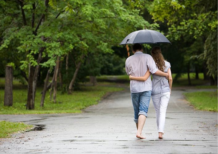 Couple walking barefoot under an umbrella on a wet path in a park.