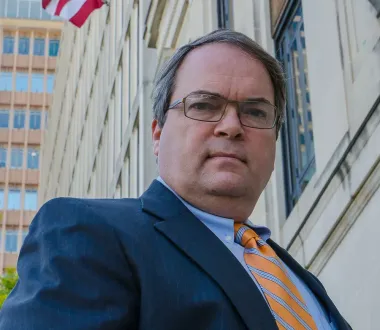 Man in navy suit and orange tie standing outside a government-style building with an American flag