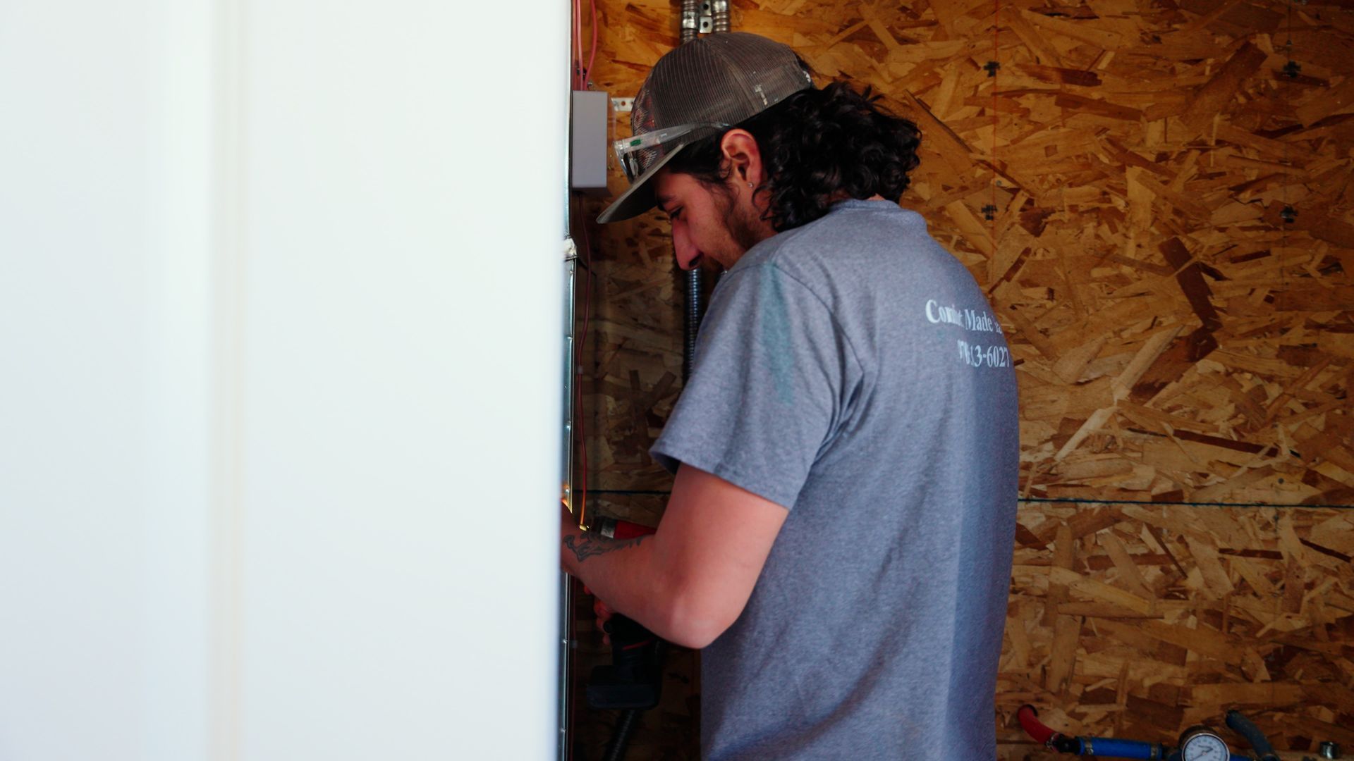 Worker in a cap and gray shirt inspecting wiring inside a wall frame.