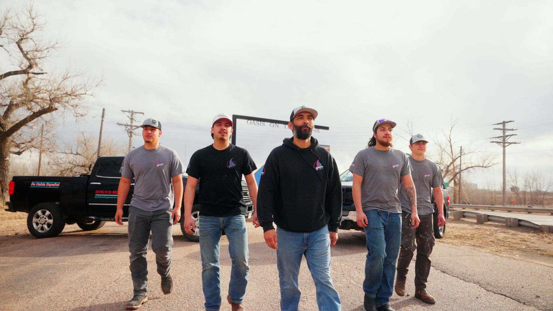 Five men walking toward the camera on a rural road beside a black pickup truck, with bare trees and utility poles.