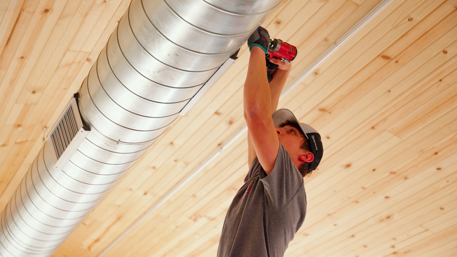 Person using a power drill to install a silver air duct on a wooden ceiling