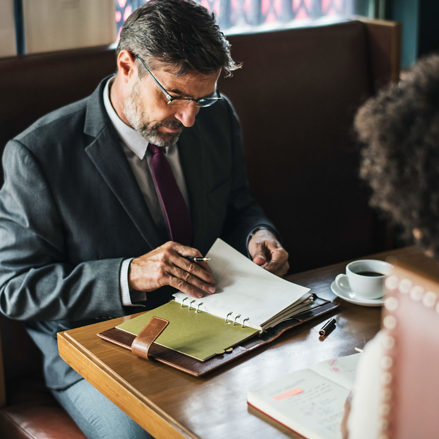 Man in a suit reviewing papers at a café table with a laptop and coffee cup