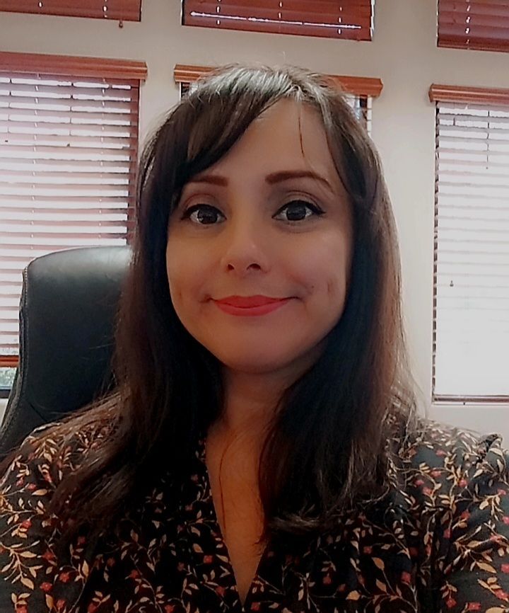 Woman smiling in an office, seated before window blinds, wearing a dark patterned blouse.