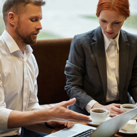 Two people in business attire discuss documents at a table with a laptop and coffee cups.
