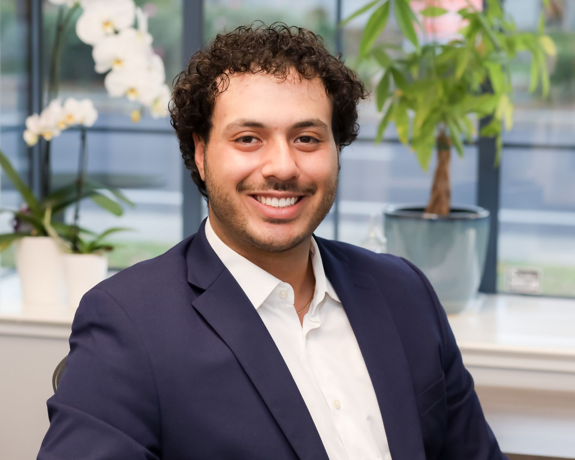 Smiling man in a navy blazer standing indoors with potted plants and flowers in the background