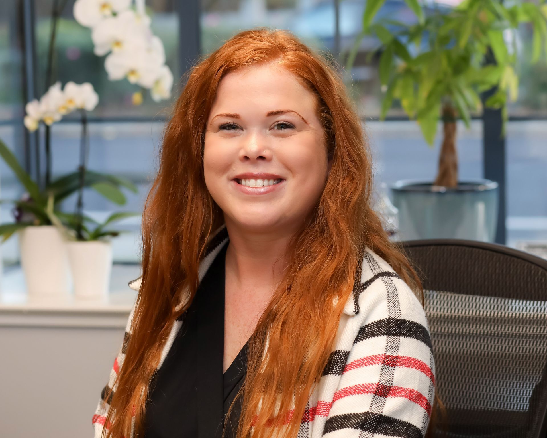 Smiling woman with long red hair seated in an office, with flowers and plants in the background
