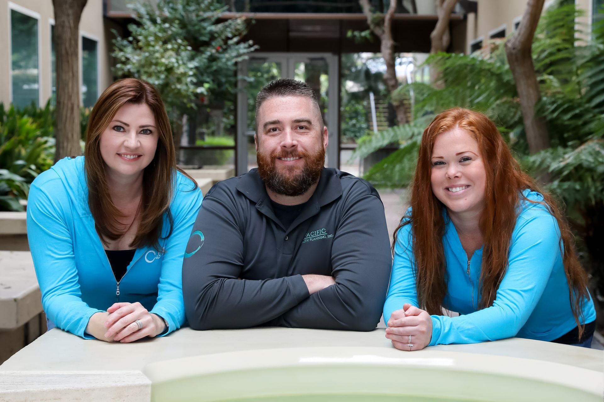 Three smiling people lean on a table in a bright indoor courtyard with plants.