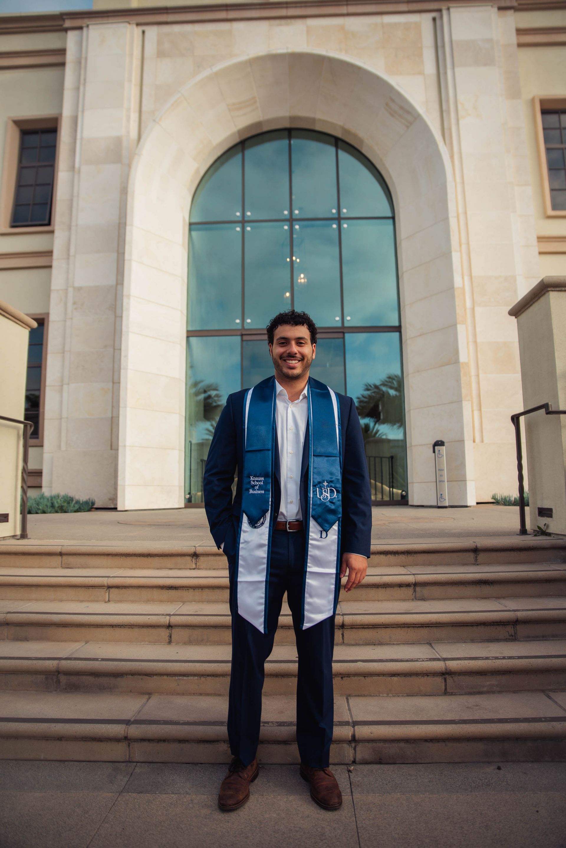 Person standing on steps in front of a large arched glass doorway, wearing dark clothes and a blue scarf.